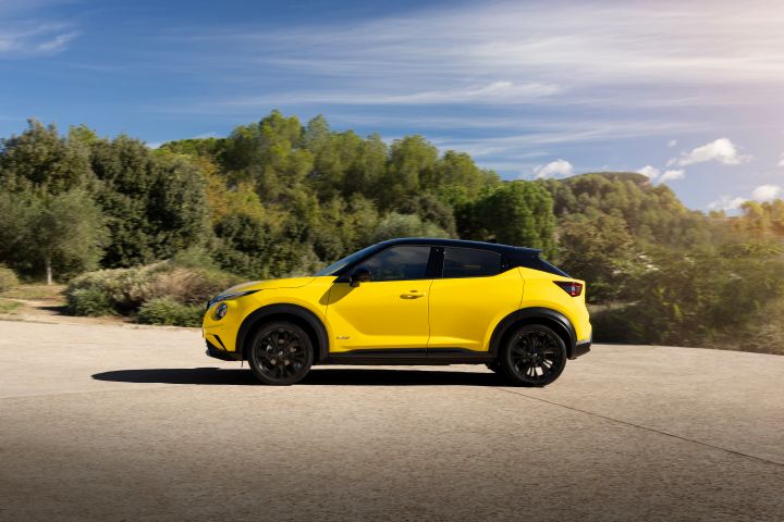 Seitliche Fahraufnahme gelber Nissan Juke auf hellem Untergrund vor grünen Bäumen und Büschen und blauem Himmel