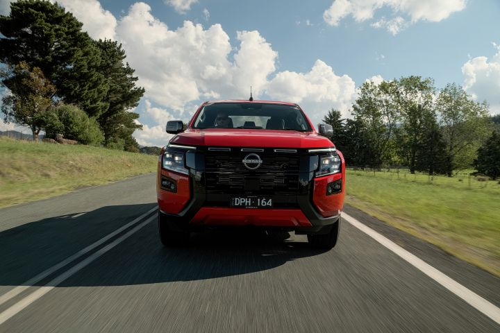Front‑facing motion shot of a 2026 Nissan Navara ST‑X Outback Red dual cab on a sealed rural road lined with grass and scattered trees.