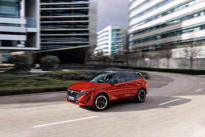 Nieuwe Nissan Qashqai in de kleur Fuji Sunset Red, rijdend op een weg langs hoge, moderne gebouwen