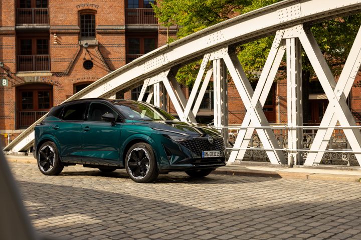 Ein Nissan Qashqai leuchtet grün in der Sonne, während das Fahrzeug auf einer gepflasterten Brücke in der Hamburger Hafenstadt vor roten Backsteingebäuden steht