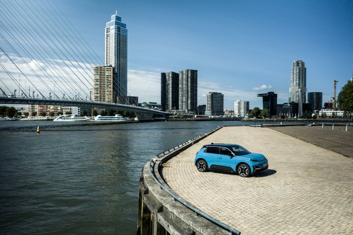 Ein blauer Nissan Micra parkt auf einem gepflasterten Platz vor der Silhouette von Rotterdam