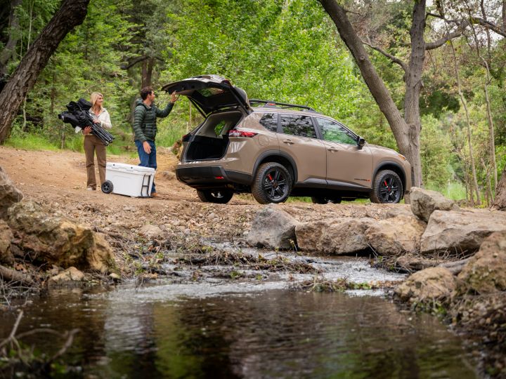 Right rear 3/4 view of the 2025 Nissan Rogue rock creek parked in a wooded area with two people grabbing items from the trunk.