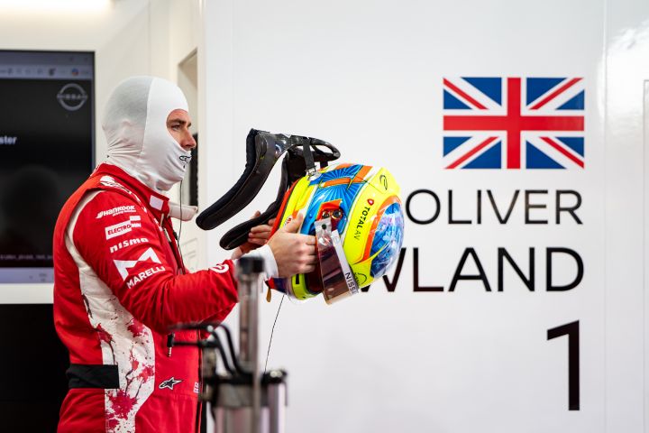 Oliver Rowland hält seinen Helm in der Teamgarage in der Hand, während er seinen roten Team-Rennanzug trägt.
