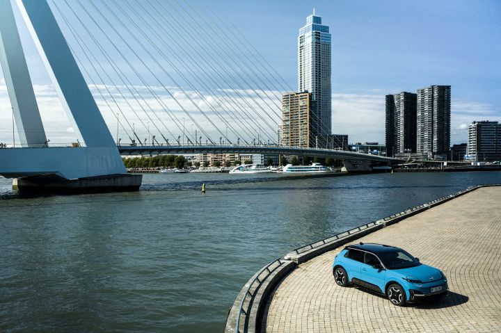 Eine große weiße Brücke spannt sich über das Wasser im Zentrum von Rotterdam, rechts hinten stehen mehrere Hochhäuser und vorne rechts parkt ein blauer Nissan Micra auf einem hell gepflasterten Untergrund
