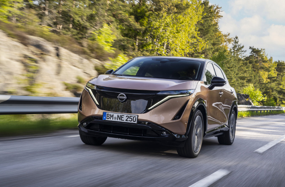 Koperkleurige Nissan ARIYA, in de kleur Akatsuki Copper, schuin links van voren, rijdend over een zonnige, bosrijke landweg.