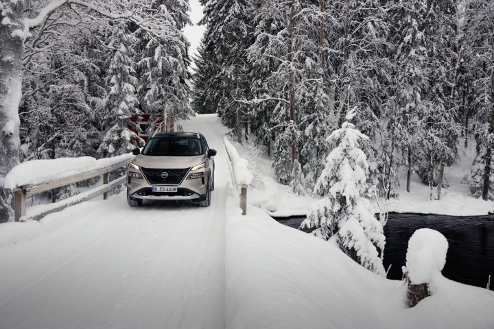 Silberner Nissan X‑Trail e‑Power e‑4ORcE fährt auf schneebedeckter Brücke mit verschneitem Wald im Hintergrund, Front