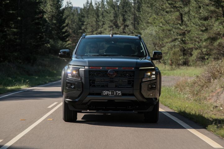 2026 Nissan Navara PRO-4X finished in Boulder Grey shown in a symmetrical frontal driving image on a straight forest road with illuminated lamps and tall evergreens behind.