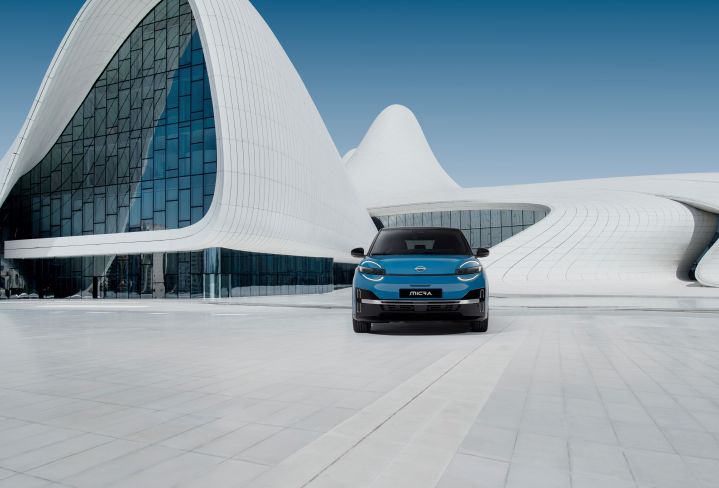 full front low view of new Micra in gear blue color and black roof positioned in front of white and blue design building with signature headlamps on and MICRA nameplate