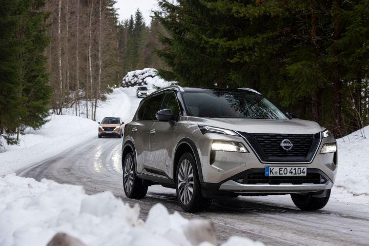Silberner Nissan X‑Trail e‑Power e‑4ORcE fährt auf nasser Fahrbahn in verschneiter Landschaft vor einem vollelektrischen Nissan Ariya