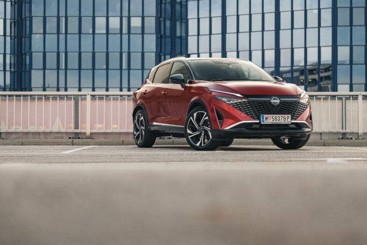 Blick von schräg vorne auf einen stehenden Nissan Qashqai in Fuji Sunset Red auf einem Parkplatz vor einem verglasten Gebäude