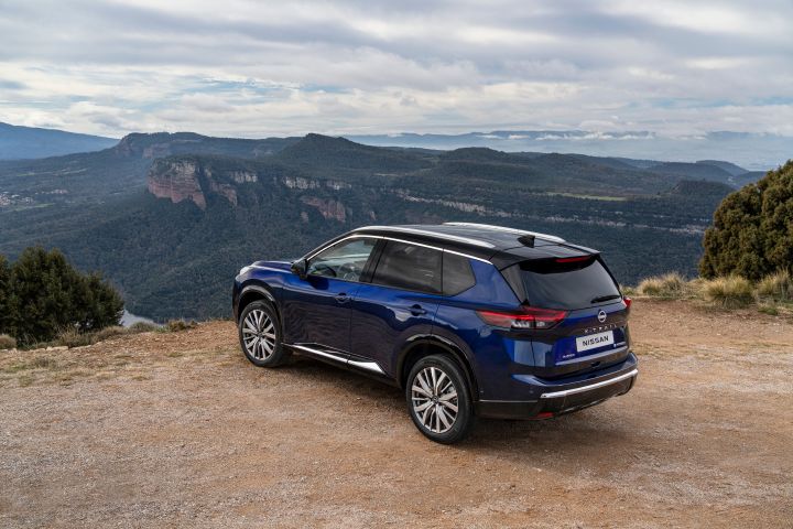 Nissan X‑Trail Tekna in der universellen Karosseriefarbe Blau, geparkt auf einem unbefestigten Aussichtspunkt mit einer bergigen Landschaft und bewölktem Himmel im Hintergrund. Das Heck und die Seitenprofile des Fahrzeugs sind sichtbar und unterstreichen sein schlankes Design, die Leichtmetallfelgen und die Dachreling.