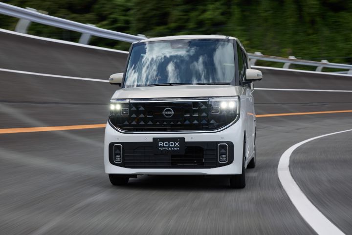 The new Nissan Roox kei car, in white with headlights on, is driving forward on a curved multi-lane road. The Nissan logo is visible on the front grille, and the road is surrounded by greenery with guardrails along the sides.