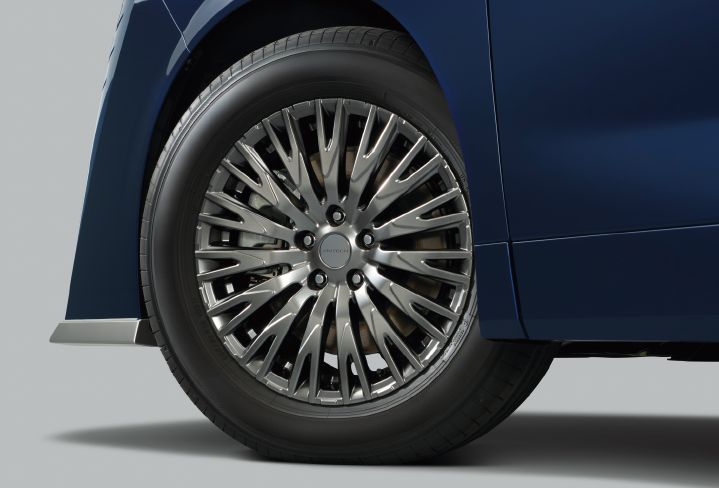 ブルーのエルグランド AUTECHのホイール周辺を、車両側面からクローズアップで撮影した画像。車体は右方向を向いており、ブルーのボディ下部とホイールアーチの一部が見えている。画面中央には大径の専用アルミホイールが配置され、細く伸びた多数のスポークが放射状に広がる立体的なデザインを採用している。ホイールセンターには「AUTECH」のロゴが刻まれている。
タイヤはブラックカラーで、トレッドパターンやサイドウォールのディテールが鮮明に確認できる。ホイールの奥にはブレーキディスクやブレーキキャリパーの一部も見え、金属的な質感が際立っている。フロントバンパー下部のメタル調フィニッシュのプロテクターがサイドからも見えていて、ブルーのボディとのコントラストが上質な印象を与えている。