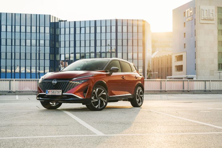Im Sonnenuntergang parkt ein Nissan Qashqai in Fuji Sunset Red vor einem verglasten Hotelgebäude