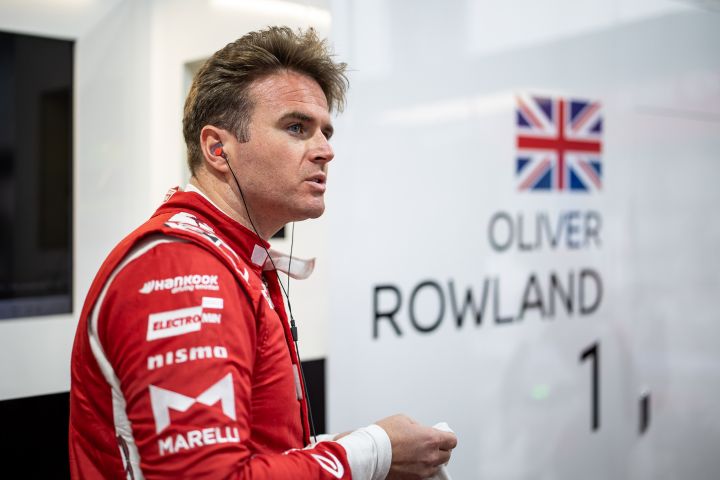 Oliver Rowland in seinem roten Rennanzug bereitet sich auf eine Session vor, hinter ihm ist sein Name an der Garagenwand zu sehen.