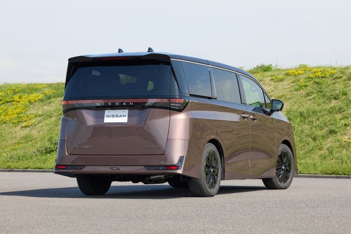 新型エルグランドが、右方向を向いた後方三分の一アングルで静止している。ブラックルーフと一体化したリアウインドウ、横一文字に広がるLEDテールランプ、中央の「NISSAN」ロゴが強調されている。背景には緑の草地と黄色い花が広がる土手があり、上空には淡い青空が広がる明るい屋外シーンで、路面は灰色の舗装となっている。
