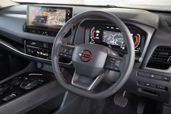 Angled view of the Nissan X‑Trail interior, highlighting the multifunction steering wheel, digital driver display, central touchscreen with navigation, and centre console controls.