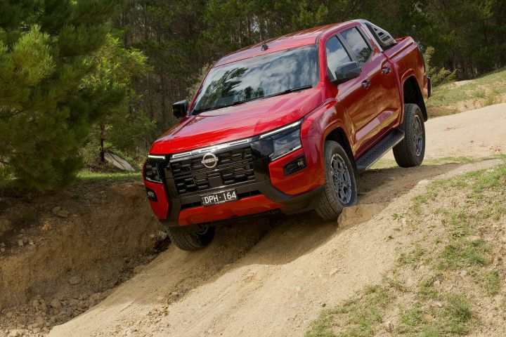 Front three-quarter view of a 2026 Nissan Navara ST-X dual cab in Outback Red, descending a steep sandy off-road track with the front wheels articulated and dense bushland behind.