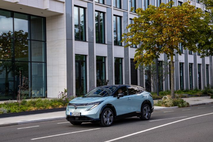 3/4 Frontansicht eines hellblauen Nissan Ariya, der auf einer Straße vor einer Bürofassade mit hohen Fenstern steht