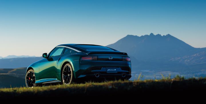 Unryu Green Fairlady Z parked on a grassy plateau, photographed from a rear-left three-quarter angle. The vehicle faces toward the left side of the image, showcasing its rear design, pronounced fenders, black alloy wheels, and dual exhaust outlets. A dramatic mountain range stretches across the background beneath a clear blue sky, highlighting the rich Unryu Green body color. The scenic natural setting emphasizes the Fairlady Z's sporty styling and strong road presence.