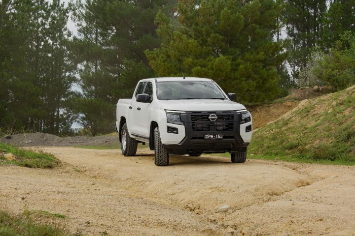 2026 Nissan Navara SL dual cab ute finished in Alpine White photographed from the front three-quarter angle on a sandy bush track, featuring the black grille, steel wheels and surrounding pine trees.