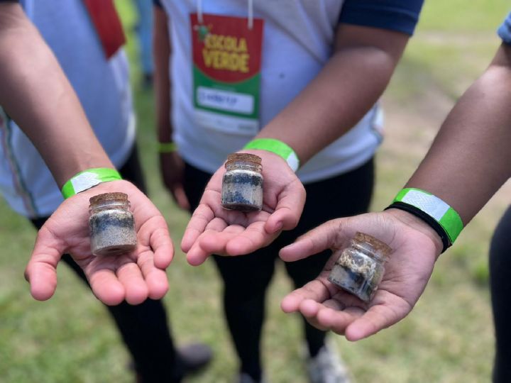 Três mãos de crianças seguram pequenos potes de vidro com terrário feito no projeto Escola Verde. Pendurado no pescoço de uma delas, um crachá mostra o logotipo do programa