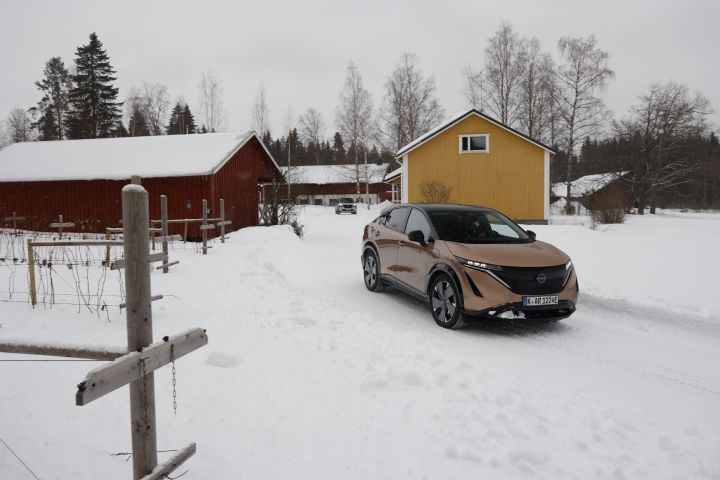 Elektroauto Nissan Ariya e‑4ORCE in Akatsuki Copper mit Dach und Außenspiegel im Schnee vor einem roten und einem gelben Holzhaus