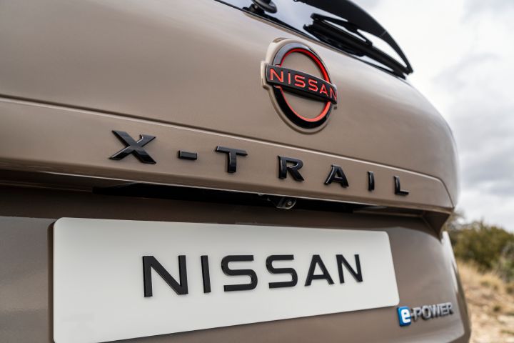 Neuer 2026 Nissan X‑Trail SUV N-Trek in der Außenfarbe Baja Storm mit Heckemblem und Typenschild. Im Mittelpunkt stehen das Nissan-Logo mit magmaroten Akzenten, der Schriftzug „X‑TRAIL“ über dem Nummernschild und das kleine blaue „e‑POWER“-Emblem, das auf die Hybridtechnologie hinweist.
