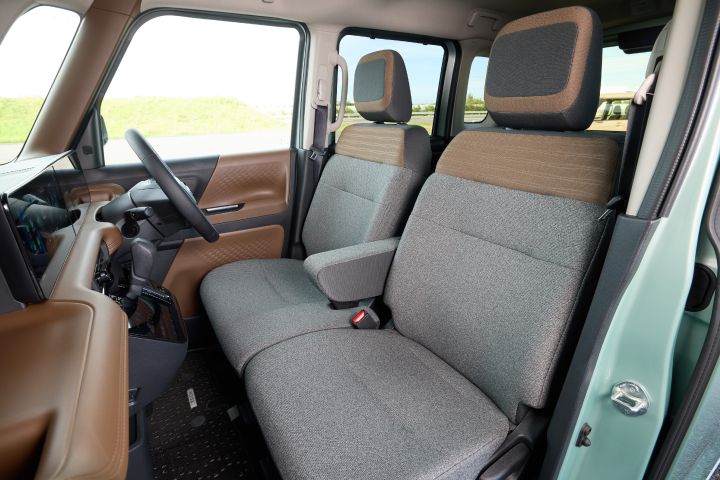 The driver and passenger seats of the new Nissan Roox kei car feature an interior color scheme of charcoal gray. The seats are uniformly charcoal gray, with brown accents on the headrests and door panels. A lush green landscape is visible through the windows.