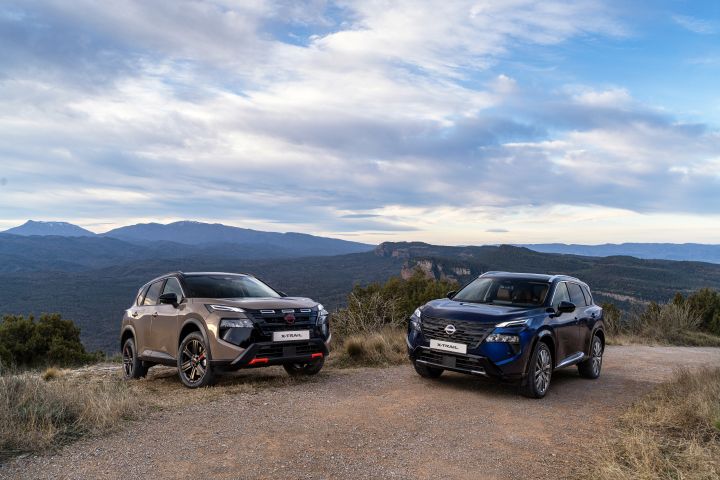 Zwei neue 2026 Nissan X‑Trail stehen auf einer unbefestigten Straße vor einer Bergkulisse und bewölktem Himmel. Die Fahrzeuge zeichnen sich durch ein modernes Design aus und sind in den Außenfarben „Baja Storm” und „Universal Blue” lackiert, die ihre robuste Geländegängigkeit und ihren Stil unterstreichen.