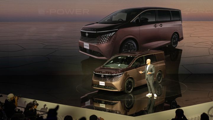 On the stage, the all-new Elgrand in Fuji Dawn and Shigoku two-tone colors is positioned diagonally, facing slightly to the left. Next to the vehicle stands CEO Ivan Espinosa in a suit, gesturing with one hand while delivering a presentation. The background features a large screen displaying a visual of the same vehicle with a clean design. In the foreground, members of the press are gathered, taking photos with cameras.