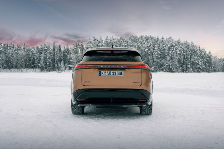 Elektroauto Nissan Ariya e‑4ORCE in Akatsuki Copper mit Dach und Außenspiegel in Pearl Black auf Eissee mit schneebedeckten Bäumen im Hintergrund, Heck