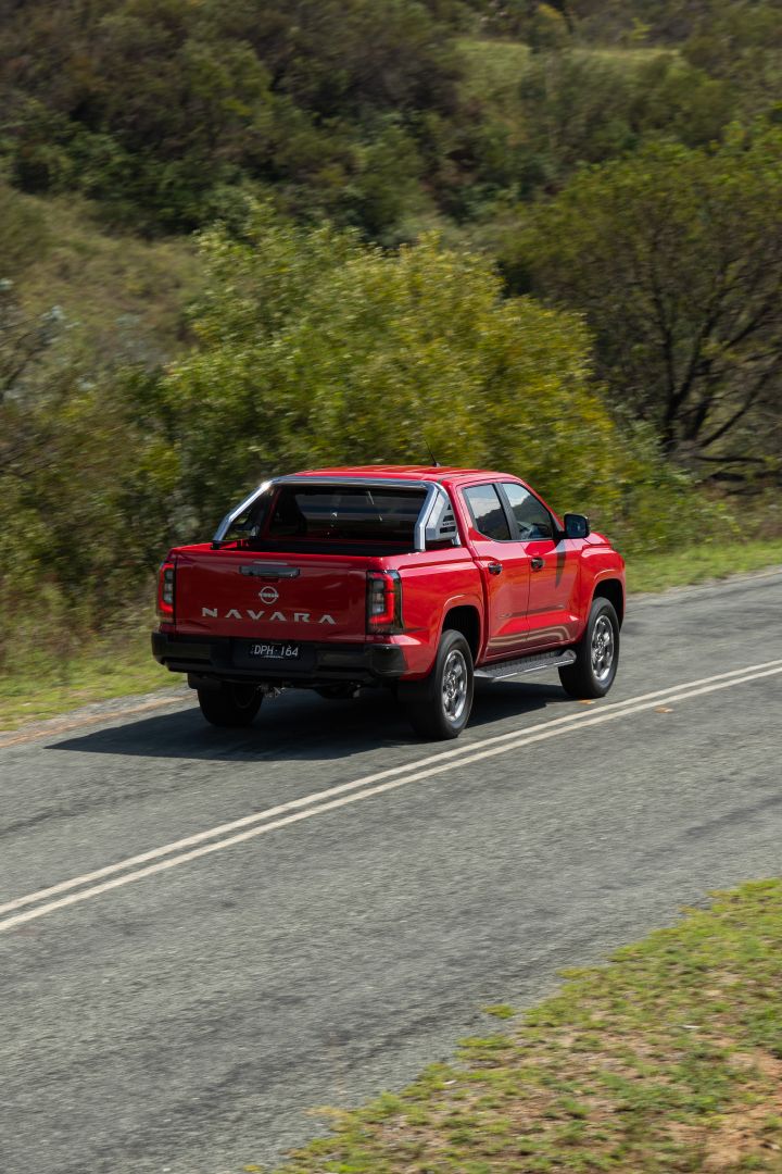 Rear three‑quarter view of a red 2026 Nissan Navara ST‑X driving on a rural road, with trees and shrubs in the background.