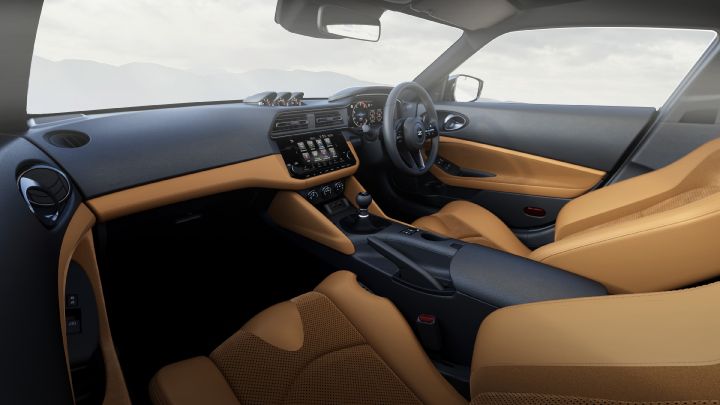 Interior view of a Fairlady Z, photographed from the passenger side facing the driver’s seat. The cabin features a two-tone design with camel brown seats and door panels, contrasted by a black dashboard and center console. A touchscreen infotainment display is positioned in the center, with physical buttons and dials below it. The driver’s area includes a round steering wheel and visible gauges. Through the windows, a soft gray sky and faint mountain silhouettes can be seen in the background, creating a calm and refined atmosphere.