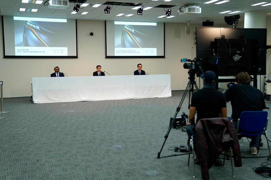 Three men sitting in a conference room in front of two large screens.