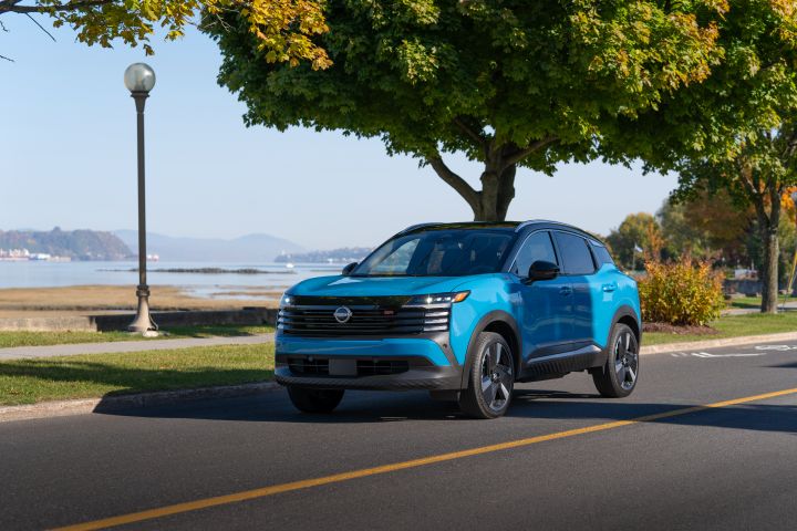 Three quarter profile of a blue 2025 Nissan Kicks parked on the road with green grass and trees in the background