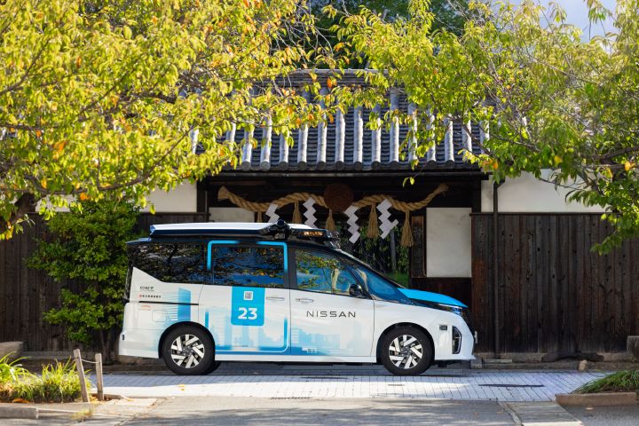 Veículo Nissan branco e azul, identificado pelo número 23, estacionado em frente a uma construção de arquitetura tradicional japonesa, cercada por árvores com folhagem verde.