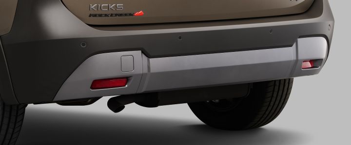 キックス「ROCK CREEK」のリヤバンパー下部を捉えたクローズアップ画像。クリスタルブラウンのボディ下部にはブラックの樹脂製バンパーが組み合わされ、その中央にはシルバーのアンダーガーニッシュが左右に広がっている。リヤゲート左側には「KICKS」と「ROCK CREEK」のエンブレムが配置され、「ROCK CREEK」ロゴにはオレンジレッドの山形アクセントがあしらわれている。