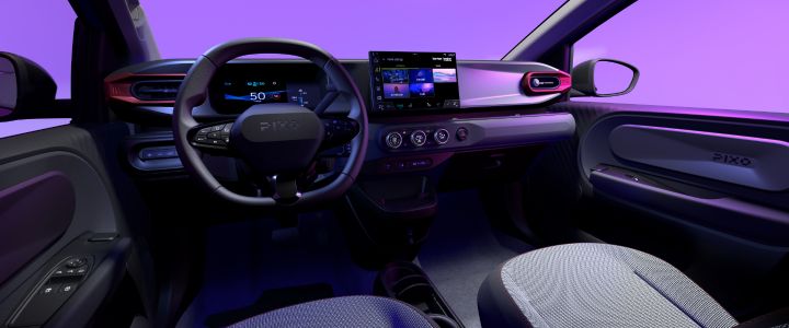 Fotografia que mostra o habitáculo de um veículo elétrico com volante, painel de instrumentos digital, ecrã tátil central e ecrã para o passageiro. A iluminação ambiente roxa, os acabamentos metálicos, os bancos com padrões e os comandos do sistema de climatização identificados criam um interior premium e de alta tecnologia.