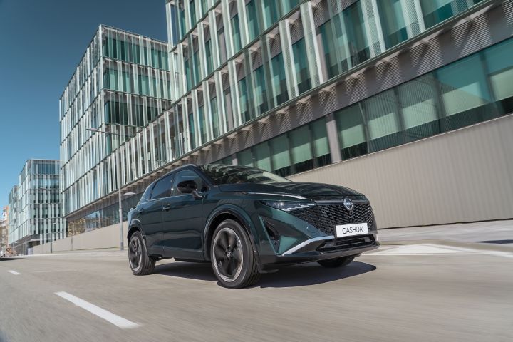 Nissan Qashqai driving on an urban road, shown from a front three-quarter angle alongside modern glass buildings