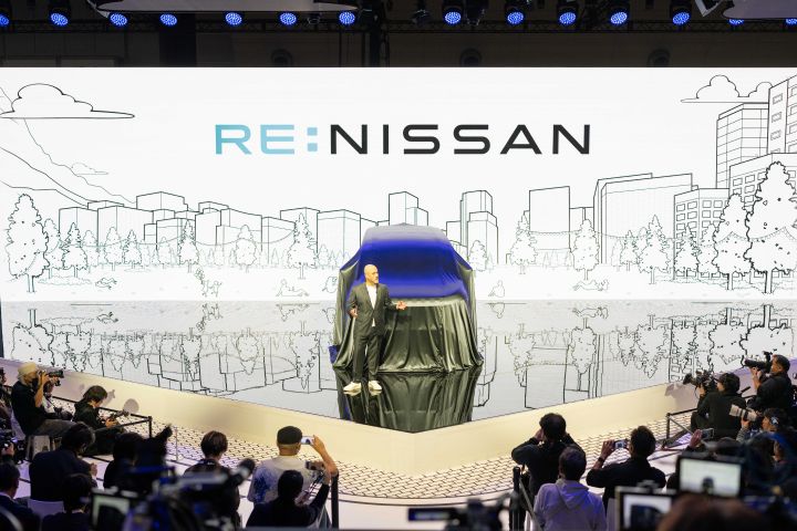 At the center of the stage, the all-new Elgrand is covered with a black cloth, and in front of it stands CEO Ivan Espinosa in a suit, gesturing with one hand while delivering a presentation. The background features a white screen with cityscape line art and the text RE:NISSAN. In the foreground, members of the press are gathered, taking photos with cameras.