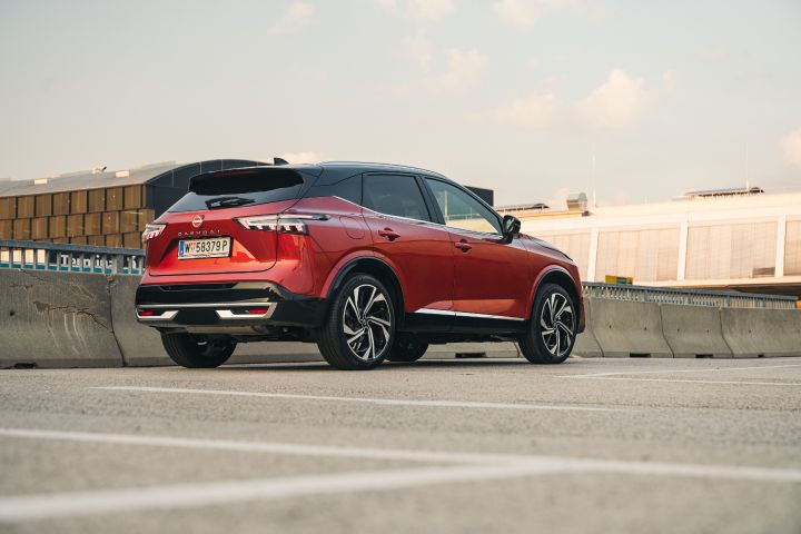 Blick von schräg hinten auf einen Nissan Qashqai in Fuji Sunset Red vor einer Industrieanlage