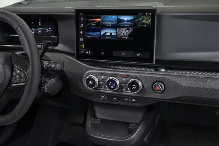 Photograph of a modern vehicle dashboard showing steering wheel, digital instrument cluster, center touchscreen, and climate-control panel. Touchscreen displays navigation, media, phone, and vehicle-function tiles, while physical knobs, USB ports, and red hazard button organize controls below.