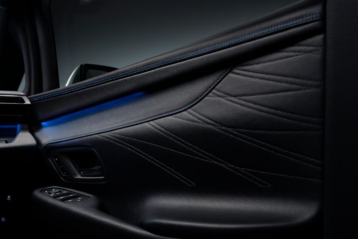 エルグランド AUTECHの運転席ドア内張りをクローズアップで撮影した画像。ブラックを基調としたドアトリムが画面全体を占め、上部から下部に向かって流れるような立体的なデザインが施されている。ドアパネルには波を思わせる曲線状のステッチパターンが広がり、細かなシボ加工が施されたレザー調素材の質感が際立っている。
上部には青色のステッチが2本通されており、その下には鮮やかなブルーのアンビエントライトが横方向に発光している。ブラックの内装と青色の照明のコントラストが、AUTECHらしい上質で先進的な雰囲気を演出している。画面左下にはブラックのドアハンドルとパワーウインドウスイッチ類が配置され、操作部周辺も落ち着いたブラックで統一されている。
