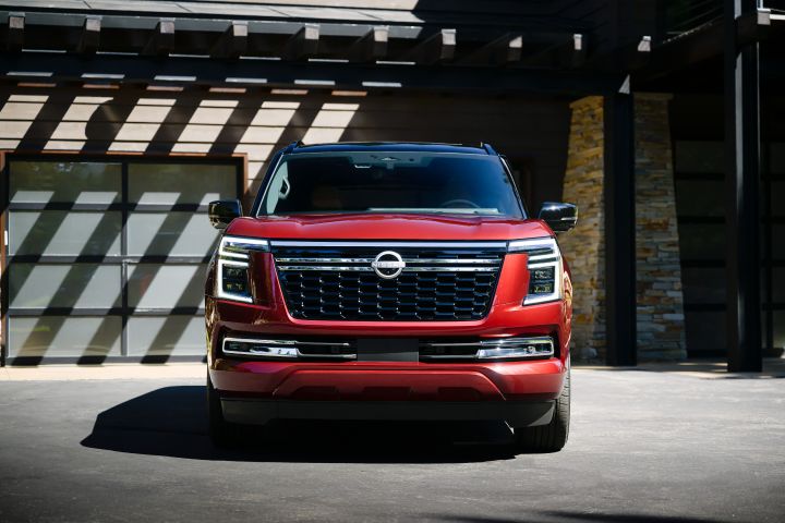 Front view of a red 2025 Nissan Armada with the headlights on parked in front of a brick building.