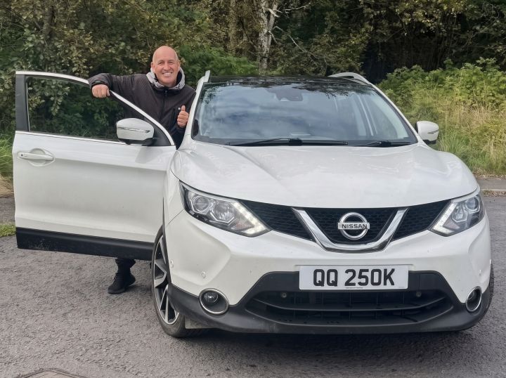 Craig Baker smiling as he stands beside with his white Nissan Qashqai with a custom number plate saying ‘QQ 250K’
