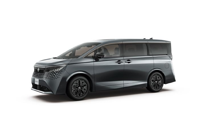 Studio image of the All-new ELGRAND in a metallic dark gray exterior color, shown from a front three-quarter view facing left. The vehicle features a black roof and dark window graphics, creating a sleek and cohesive profile. The front end is defined by a large patterned grille and slim, sharp headlamps, while the side surfaces combine smooth sculpting with strong character lines. Black alloy wheels enhance the premium and commanding appearance. The background is a plain light gray studio setting, allowing the vehicle’s design and proportions to stand out clearly.