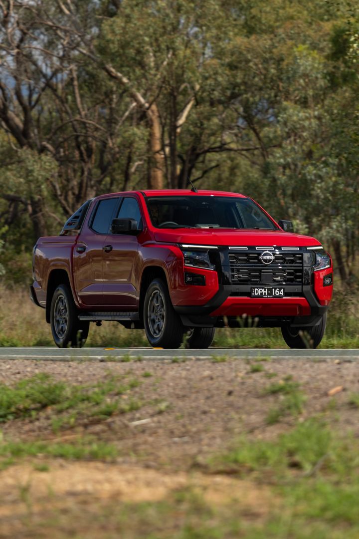 Front three‑quarter view of a 2026 Nissan Navara ST‑X dual cab in Outback Red driving on a straight highway passing through open fields and distant hills.