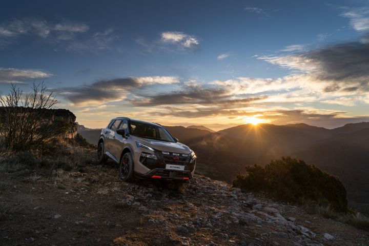 Nuevo Nissan X‑Trail of Adventure 2026 en color de carrocería Baja Storm, aparcado en una colina rocosa al atardecer, mostrando capacidad todoterreno y terreno accidentado. La escena presenta nubes dramáticas, rayos de sol rompiendo sobre montañas lejanas y vegetación natural que rodea el vehículo.