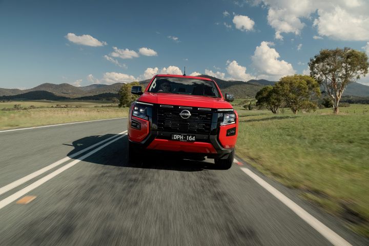Front view of a 2026 Nissan Navara ST‑X Outback Red dual cab driving on a sealed country road with hills and trees behind it.